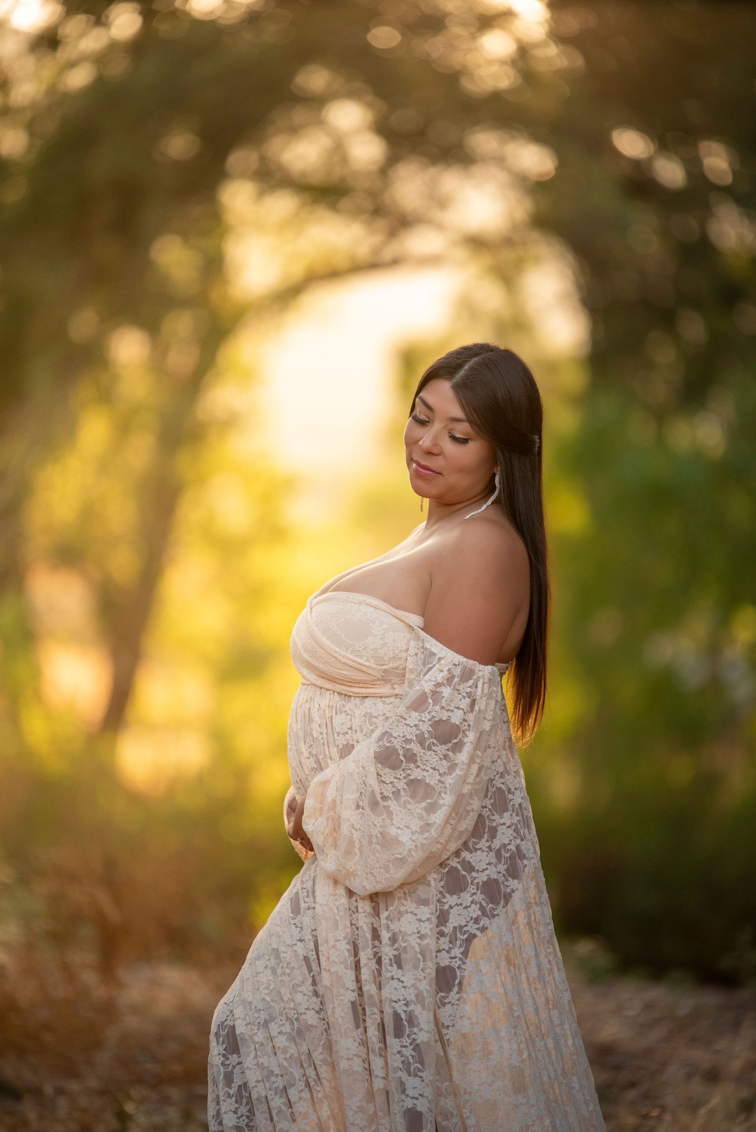 A woman in a white lace dress poses outdoors at golden hour with soft bokeh lighting in the background.