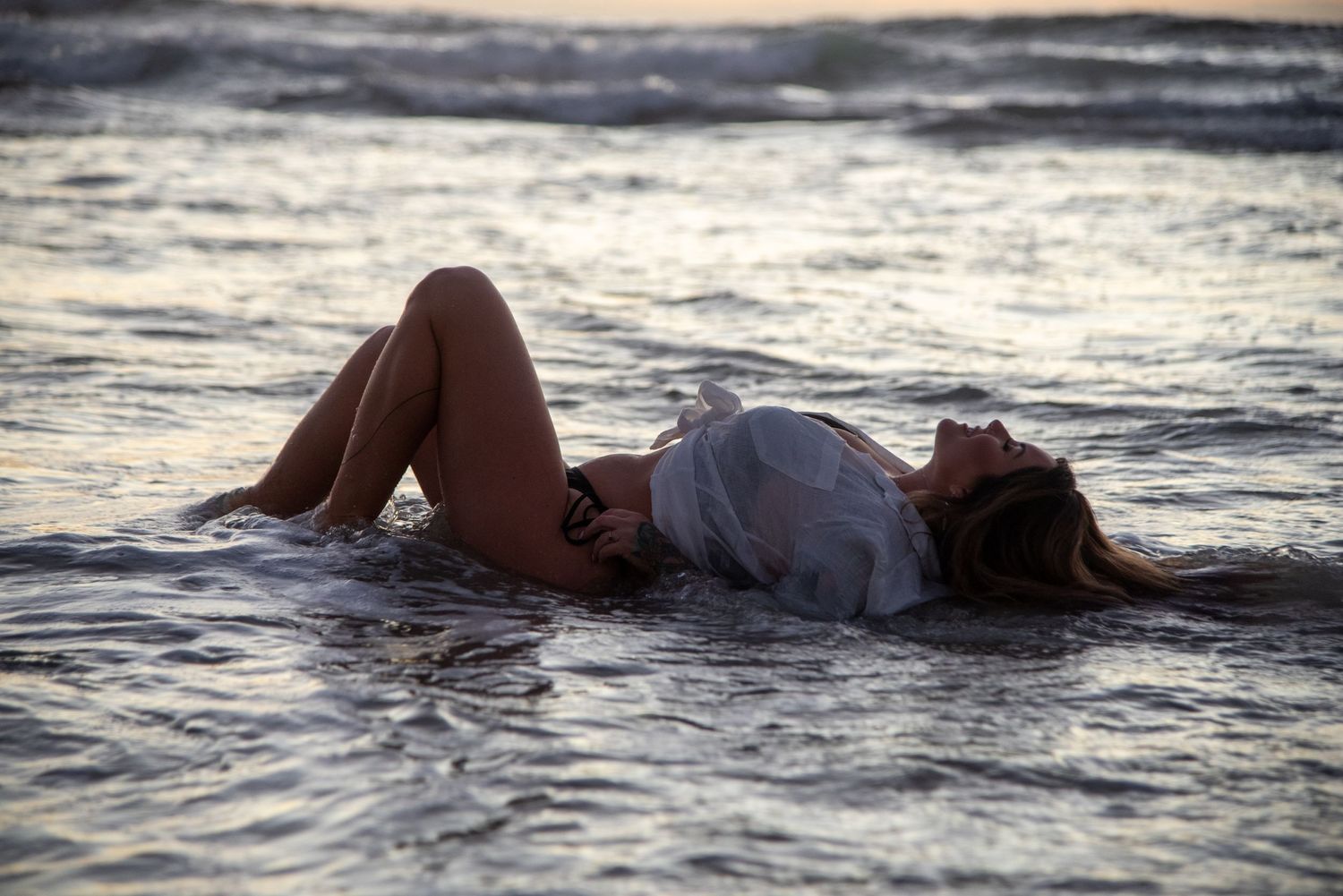 Figure in white dress laying in shallow ocean water at dusk with waves lapping around them.
