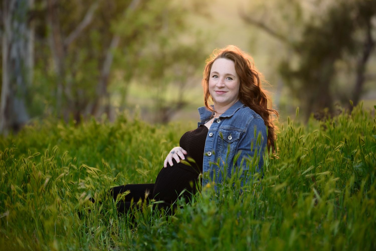 A person sits in lush green grass wearing a denim jacket during a sunny outdoor portrait session outside.