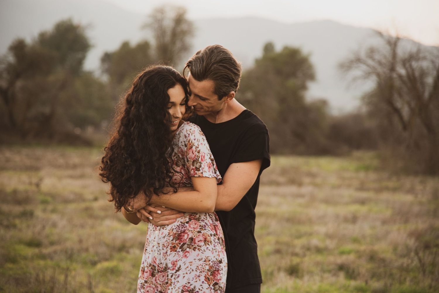 A couple embraces tenderly in a field during golden hour with trees in the background.