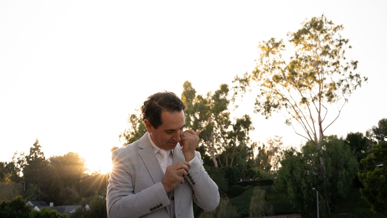 A man in a light gray suit adjusts his cufflinks outdoors at golden hour with trees and sunlight in the background.