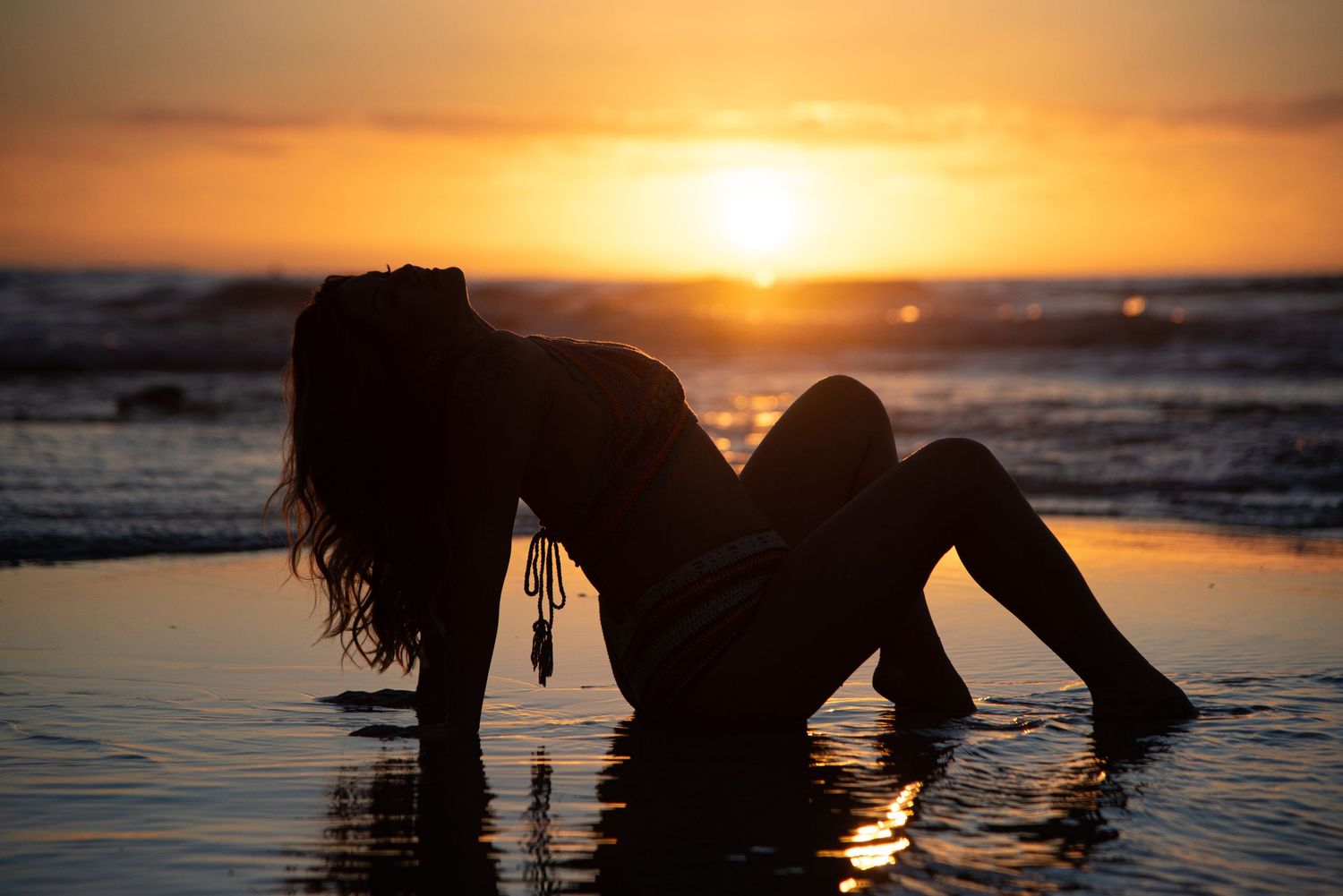 Silhouette of a figure lying in shallow ocean water at sunset with waves and golden sky in the background.