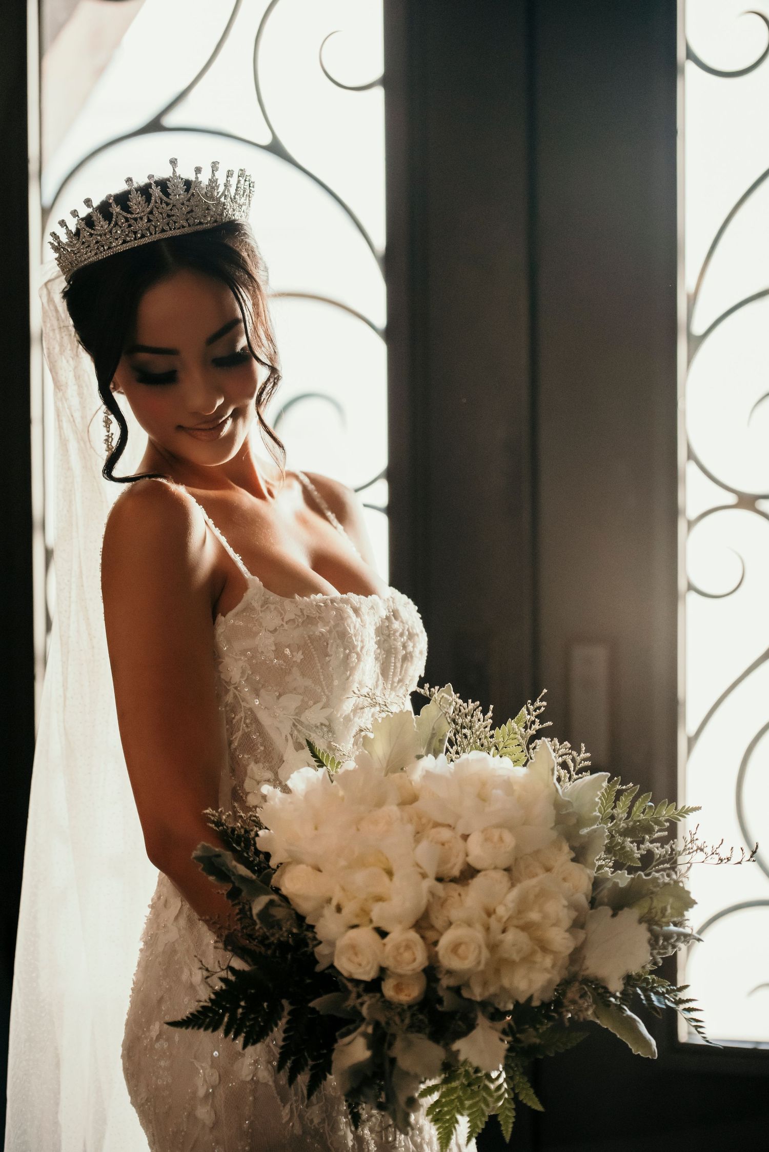 A bride in a lace gown and tiara holds an elegant white rose bouquet while standing by a decorative window.