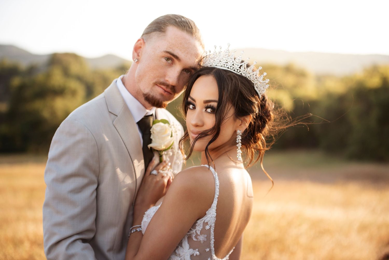 A romantic wedding couple embraces in a golden field at sunset, the bride wearing a delicate crown and holding a white rose.