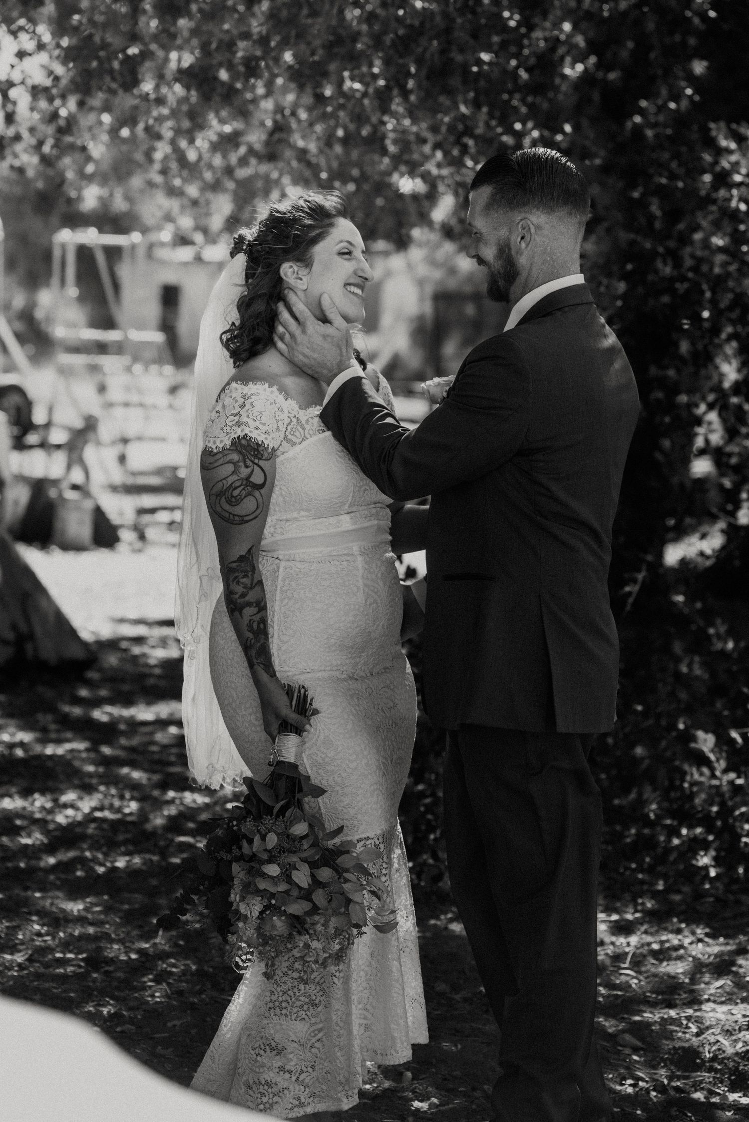 A black and white wedding moment showing a joyful couple outdoors with the bride wearing a lace dress and showing visible tattoos.