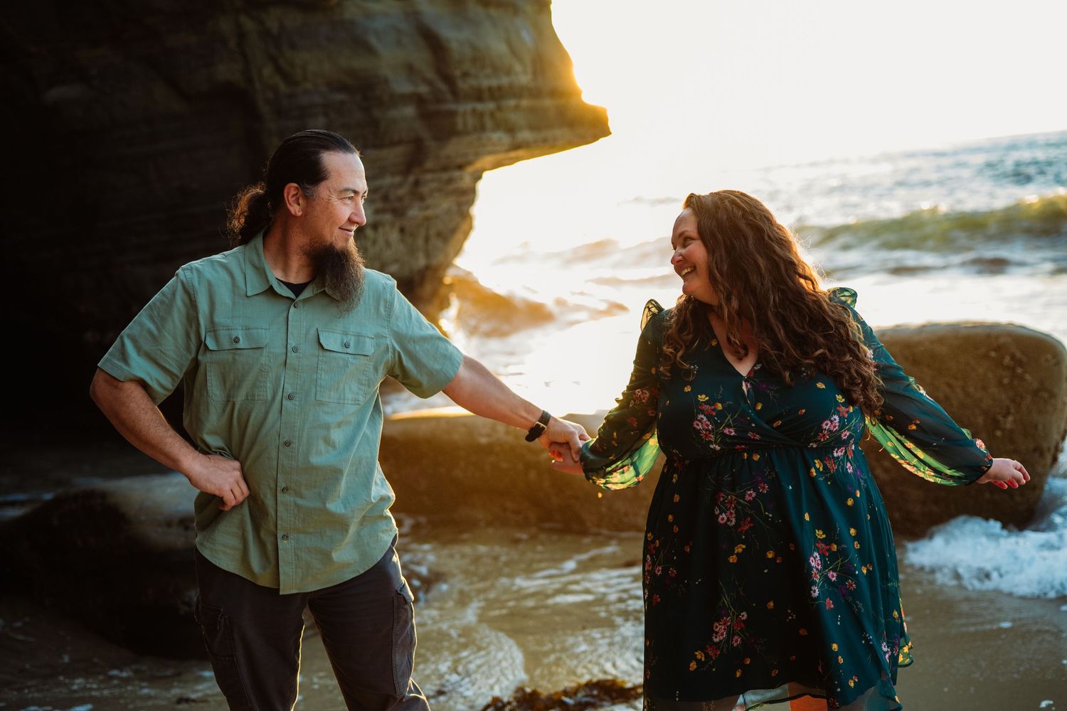 Couple holding hands and laughing on a rocky beach at golden hour with ocean waves in the background.