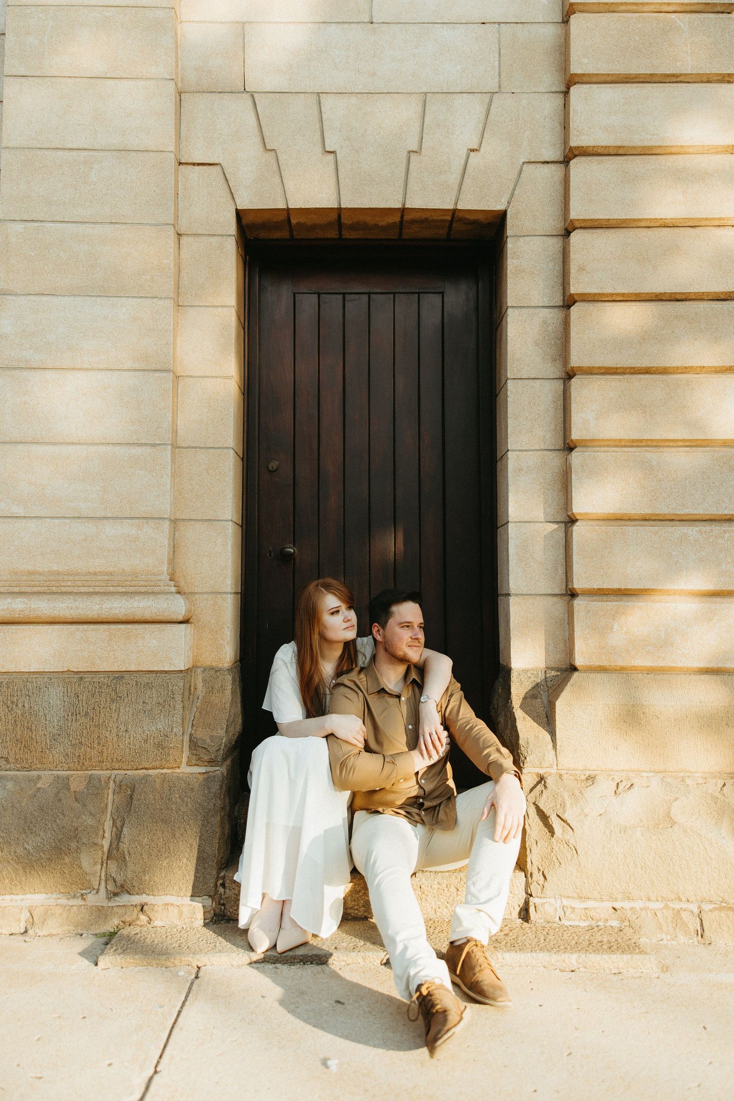 Engaged Couple sitting on a step in front of a wooden door fitted into old sandstone church building.