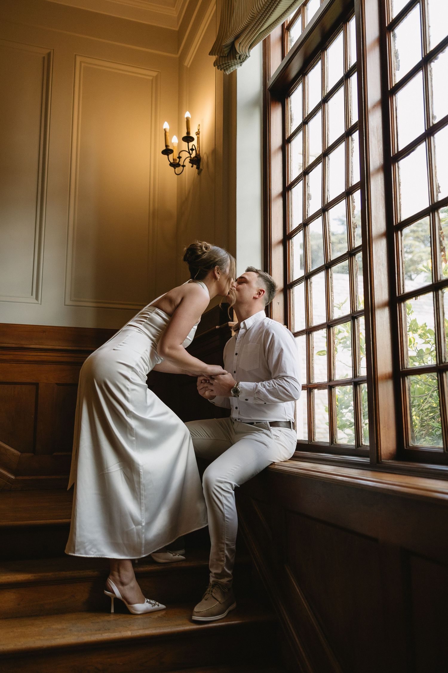 Engaged couple kissing on rich wooden stairs & wood panelled interiors of Houghton 1938, Johannesburg. She is wearing a silky white dress