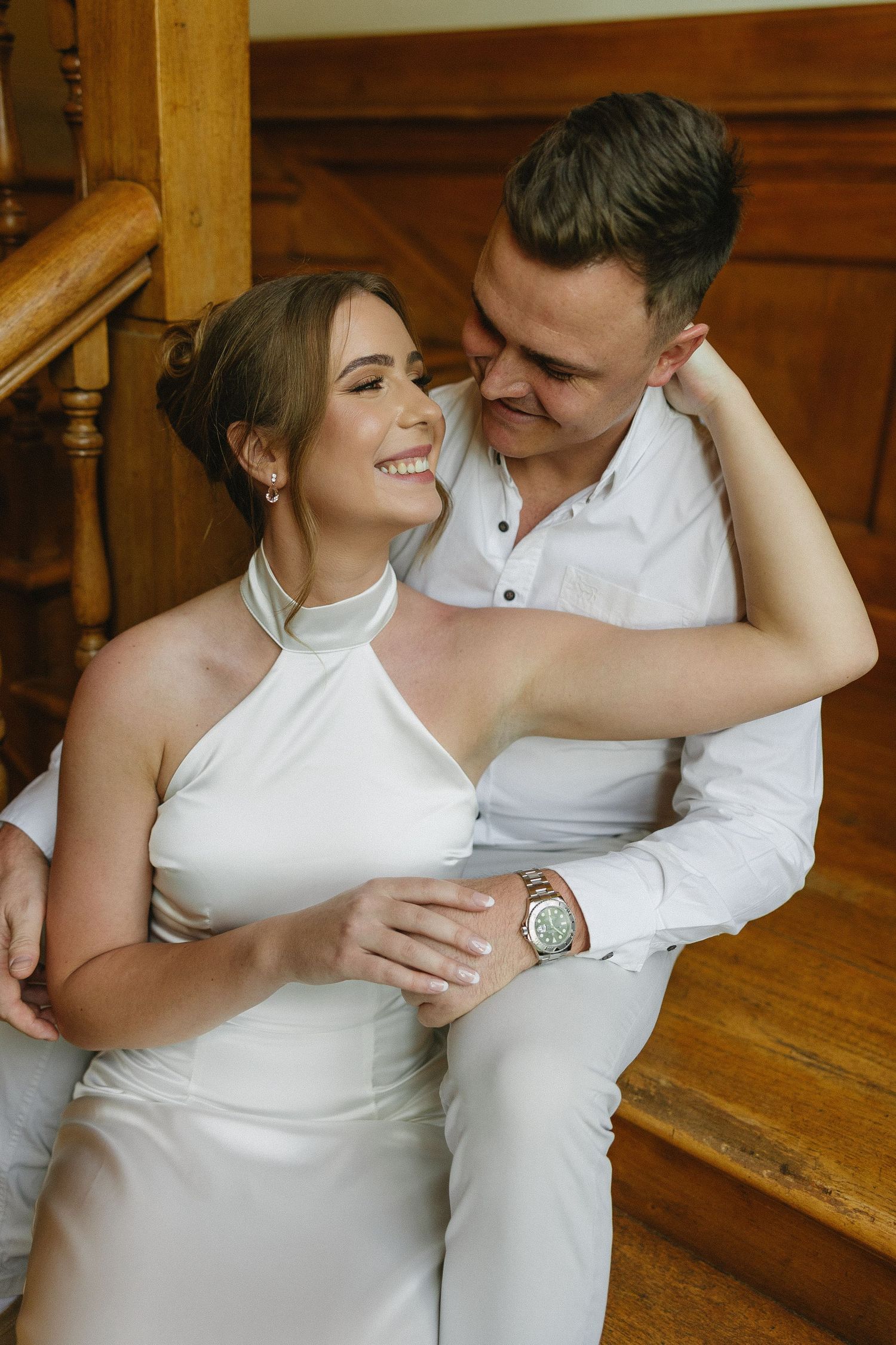 Engaged couple sitting on rich wooden stairs & wood panelled interiors of Houghton 1938, Johannesburg. She is wearing a silky white dress
