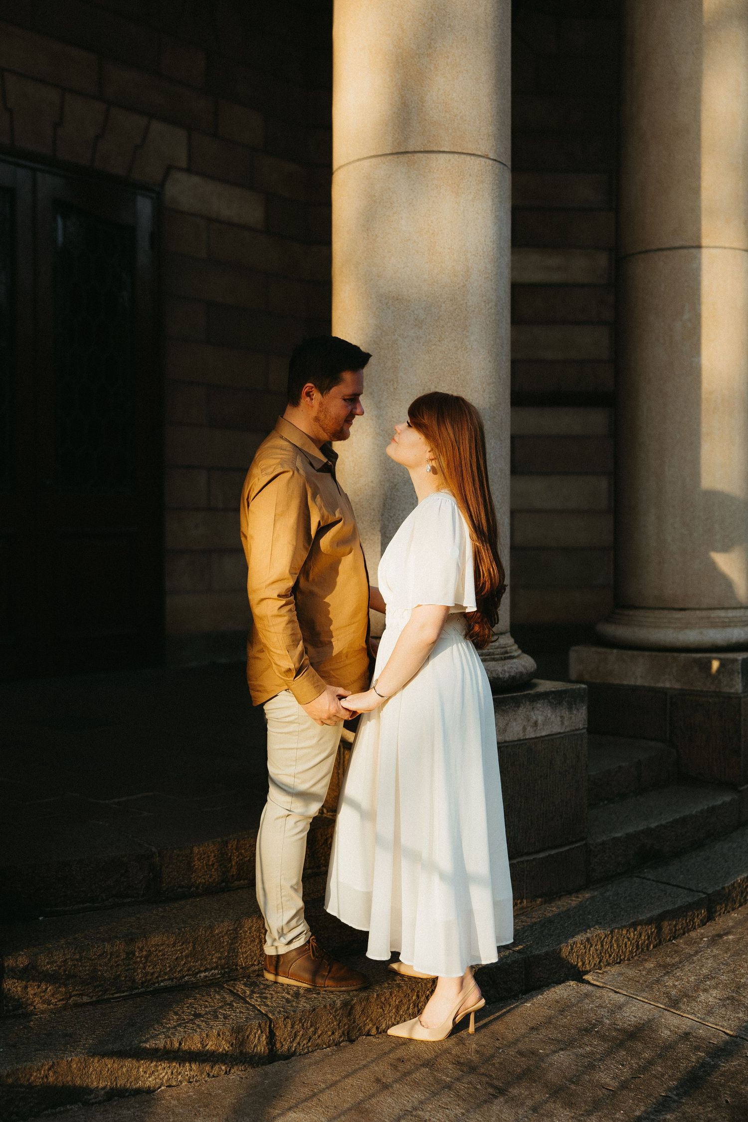 Engaged couple standing facing each other in the last bit of golden sunlight, kissing her red hair and the old sandstone church