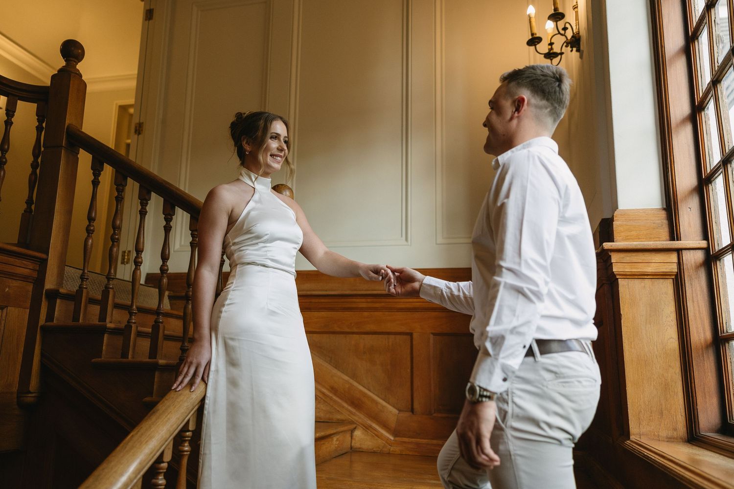 Engaged couple standing on rich wooden stairs & wood panelled interiors of Houghton 1938, Johannesburg. She is wearing a silky white dress