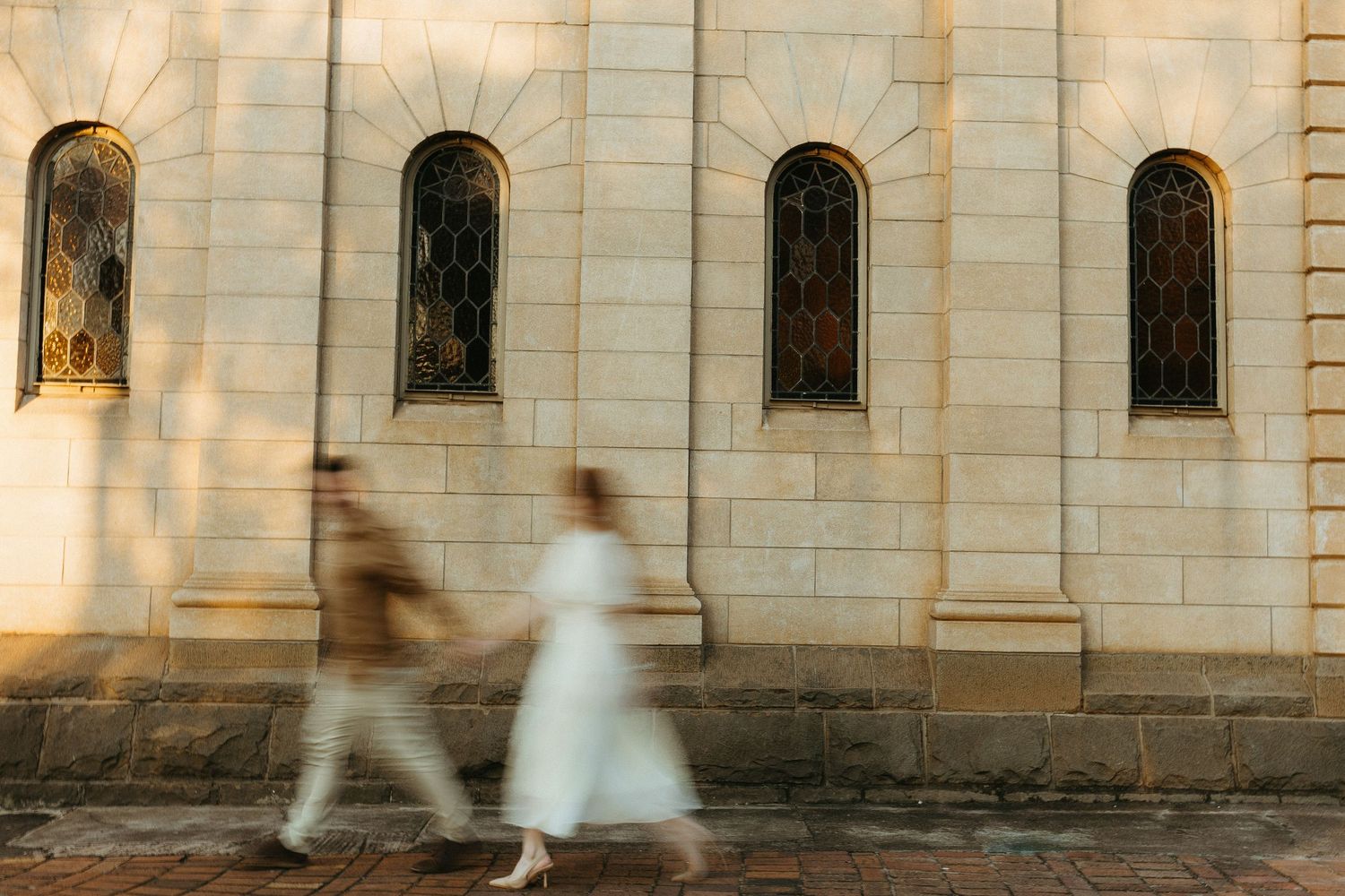 Engaged Couple walking in front of old sandstone church. The couple is blurry, capturing the movement