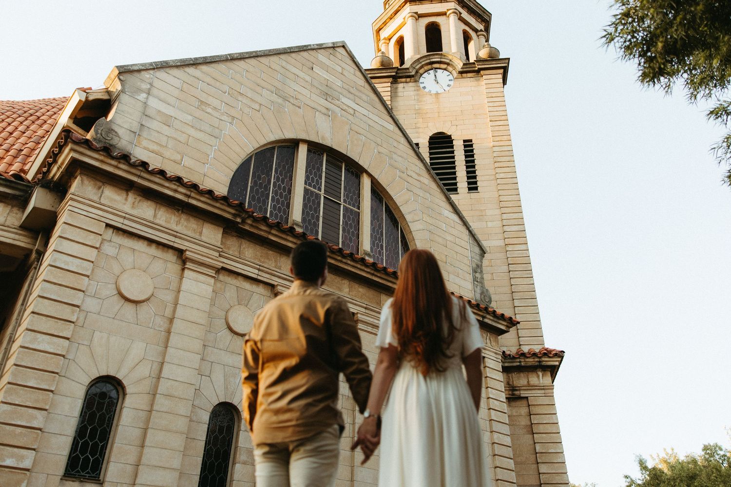 Engaged Couple facing an old sandstone church, looking at the large clock at the top