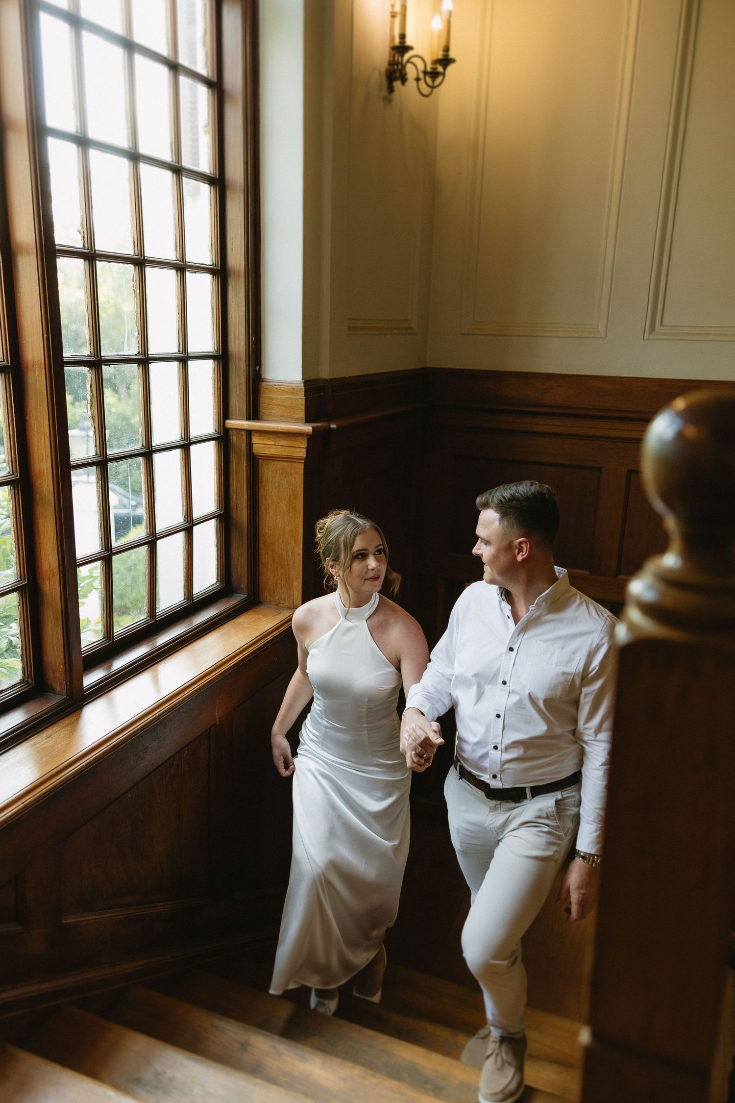 Engaged couple walking up rich wooden stairs & wood panelled interiors of Houghton 1938 in Johannesburg. She is wearing a silky white dress