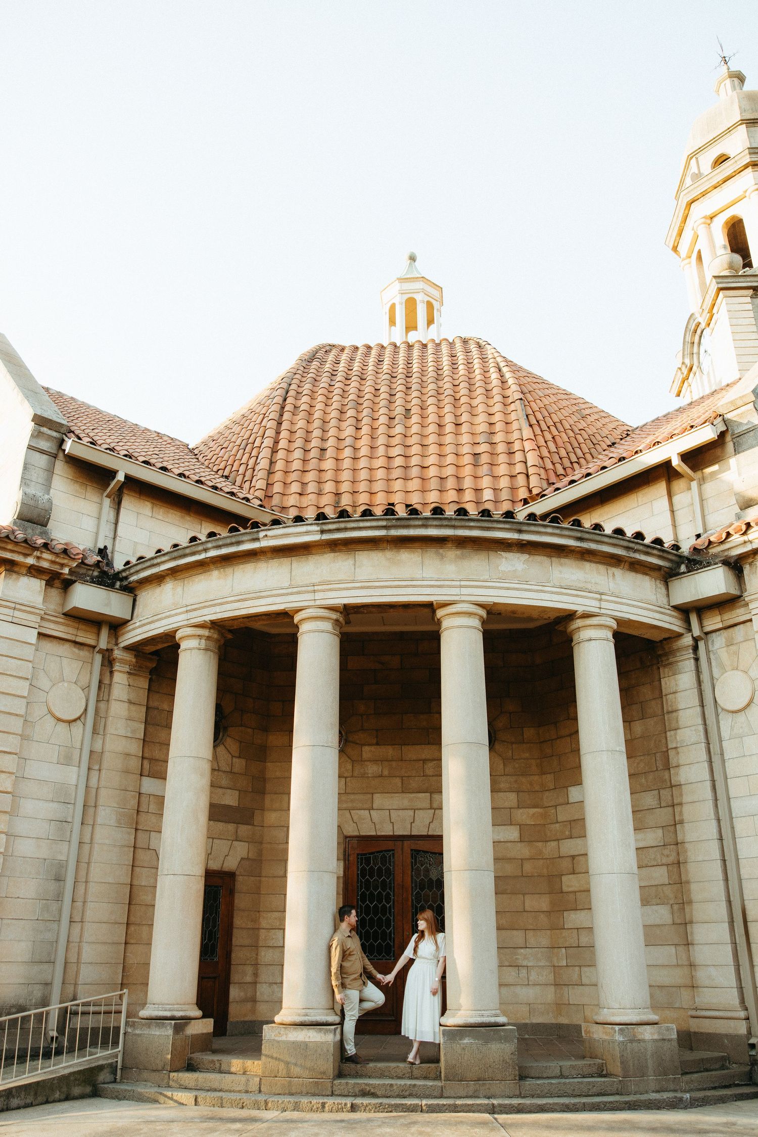Engaged Couple standing on a step in front of a wooden door fitted into old sandstone church. standing Between by 4 sandstone pillars