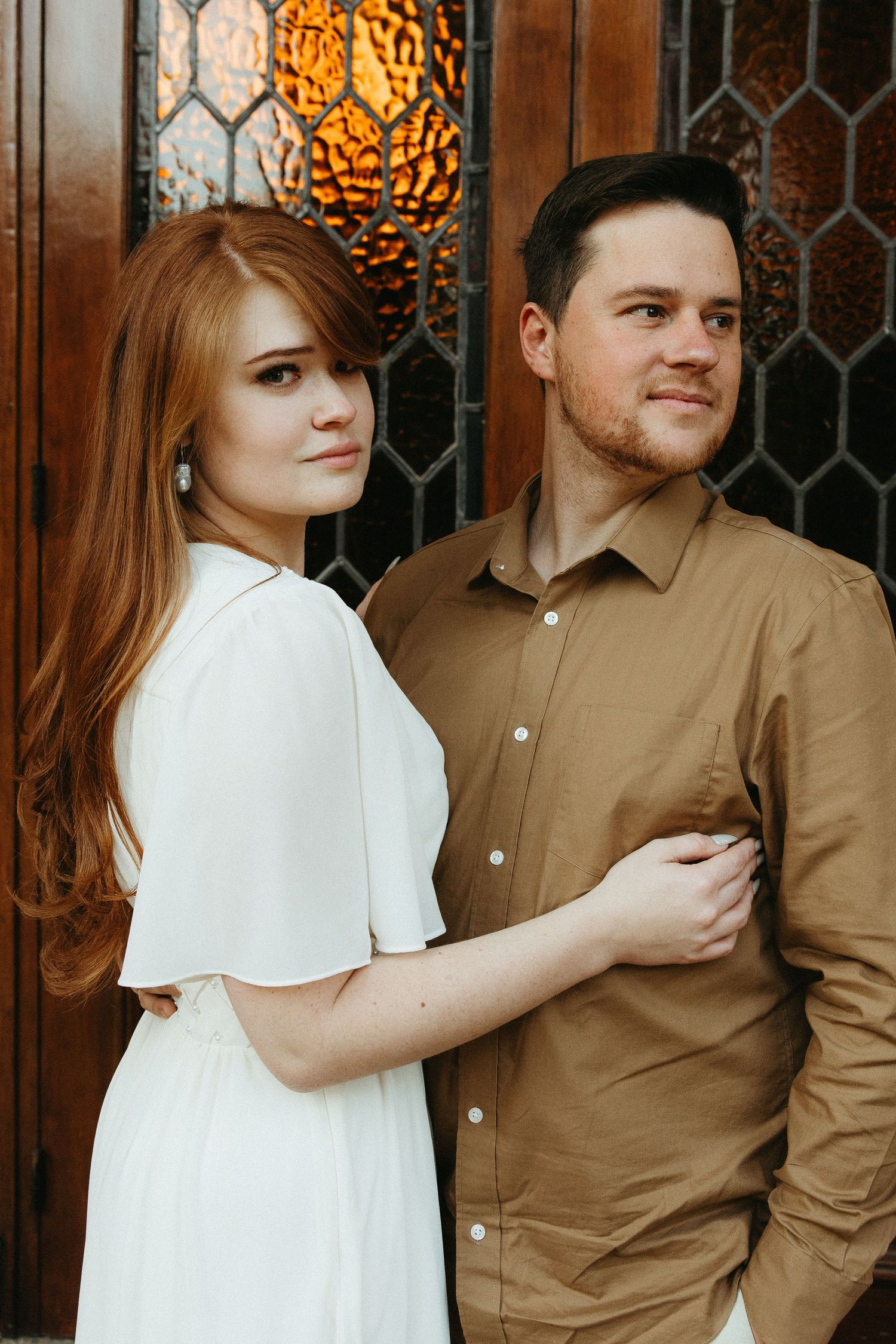 Engaged Couple standing in front of a wooden church door with orange textured glass windows.