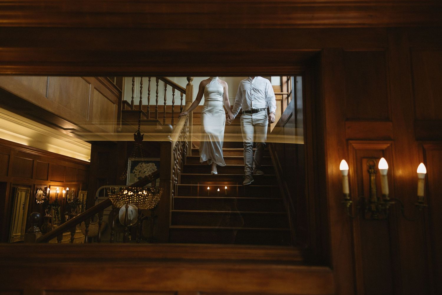 Engaged couple walking down rich wooden stairs & wood panelled interiors of Houghton 1938, Johannesburg. She is wearing a silky white dress
