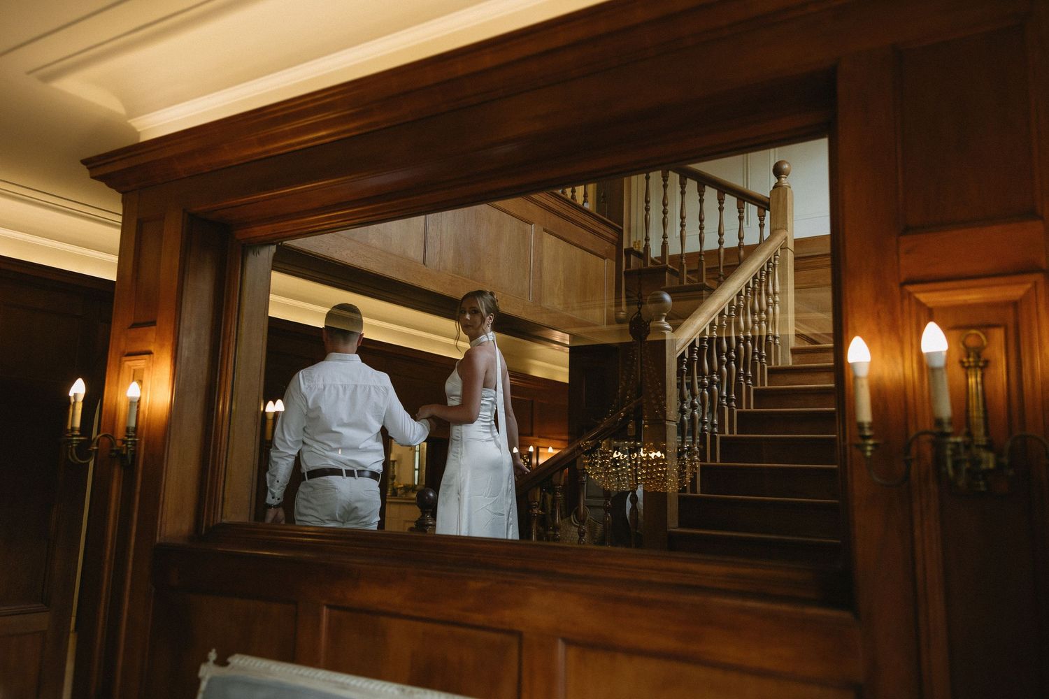 Engaged couple walking down rich wooden stairs & wood panelled interiors of Houghton 1938, Johannesburg. She is wearing a silky white dress