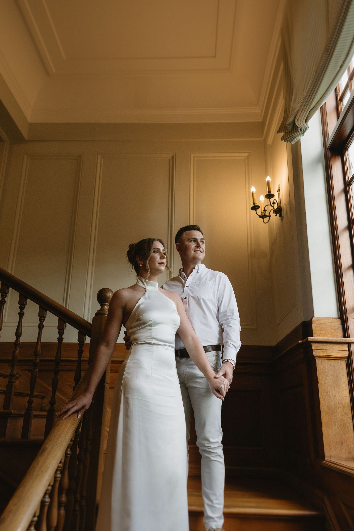 Engaged couple standing on rich wooden stairs & wood panelled interiors of Houghton 1938, Johannesburg. She is wearing a silky white dress