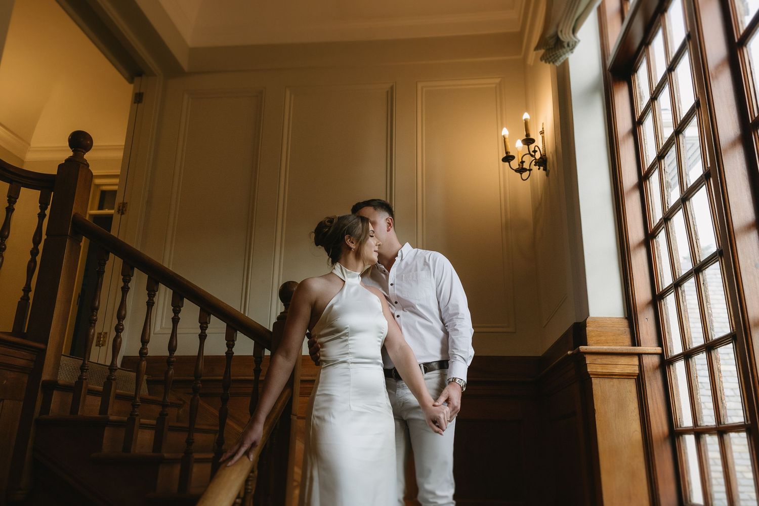 Engaged couple standing on rich wooden stairs & wood panelled interiors of Houghton 1938, Johannesburg. She is wearing a silky white dress