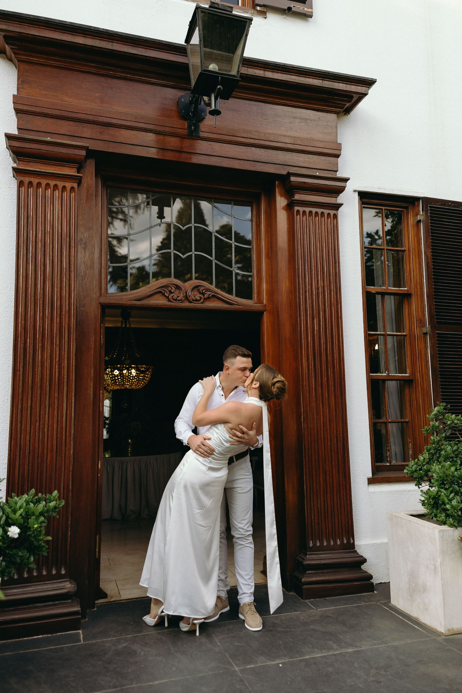 Groom-to-be dipping and kissing his Fiancé in her silky white dress in front of a stunning wooden entryway.