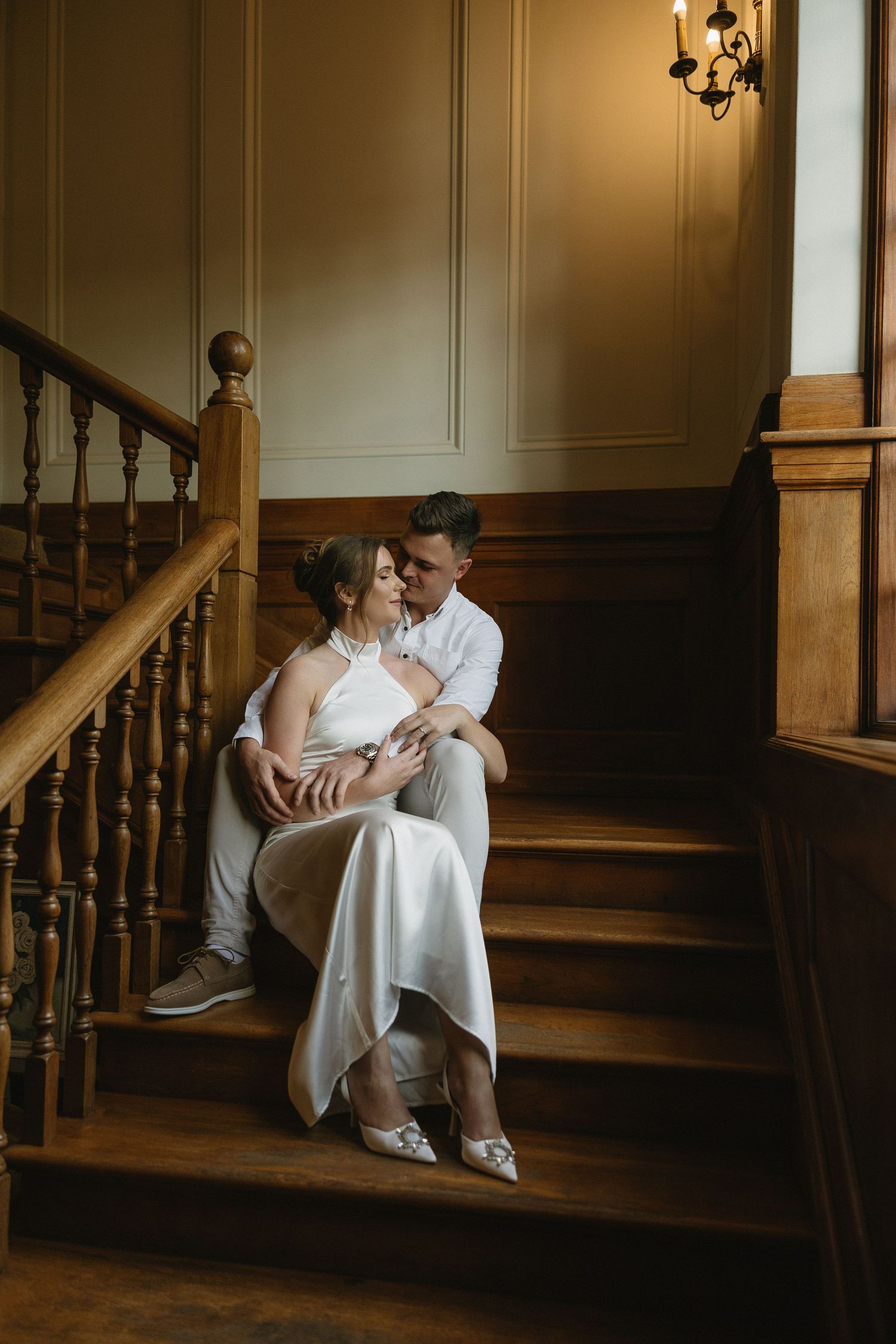 Engaged couple sitting on rich wooden stairs & wood panelled interiors of Houghton 1938, Johannesburg. She is wearing a silky white dress
