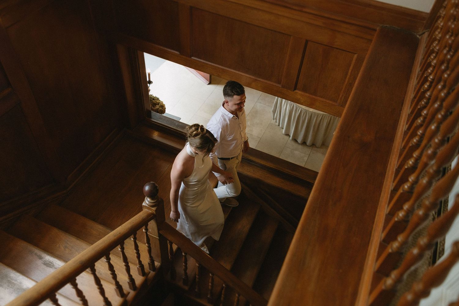 Engaged couple walking down rich wooden stairs & wood panelled interiors of Houghton 1938, Johannesburg. She is wearing a silky white dress
