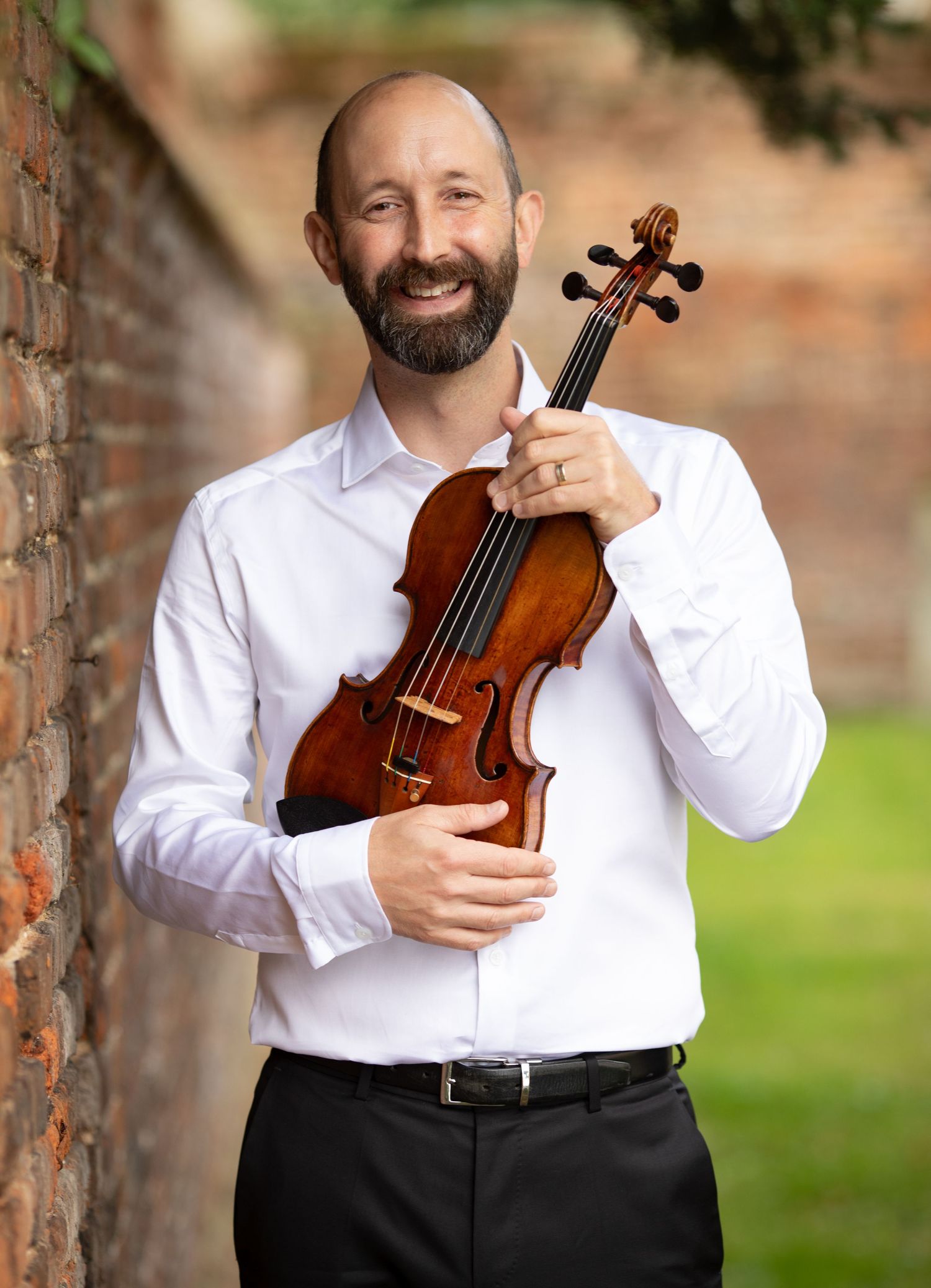 Professional musician in white dress shirt poses with violin against brick wall and garden backdrop.
