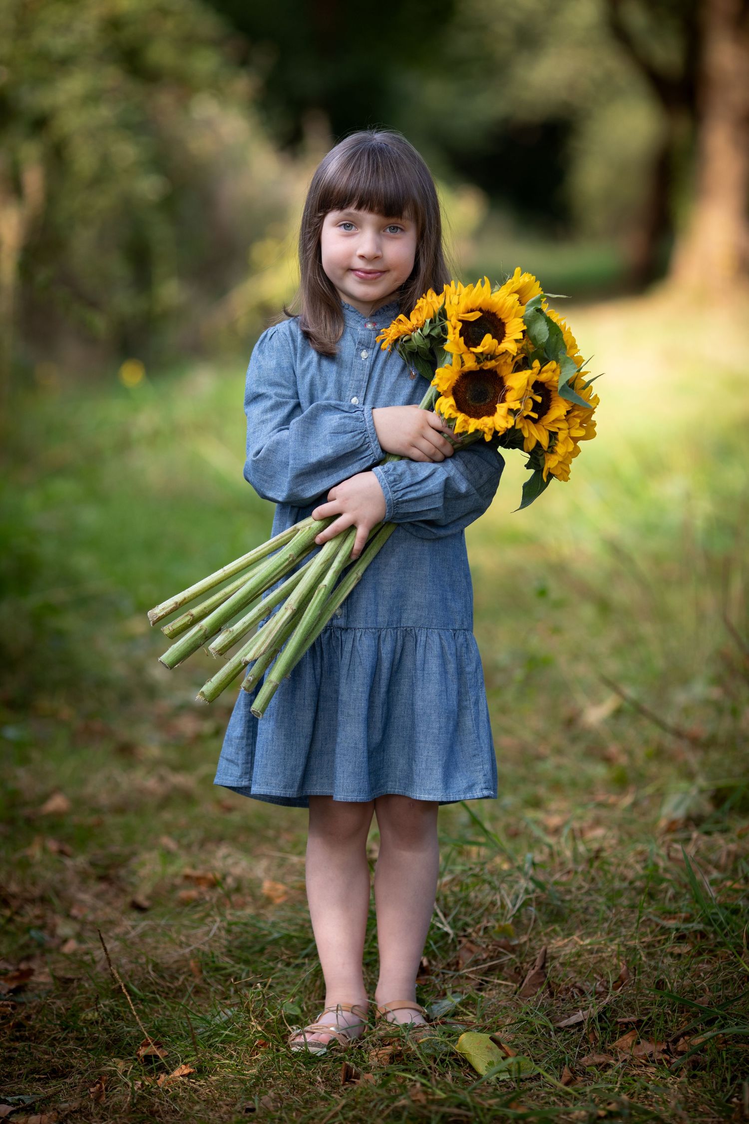 Small child in denim dress holding bright yellow sunflowers outdoors.