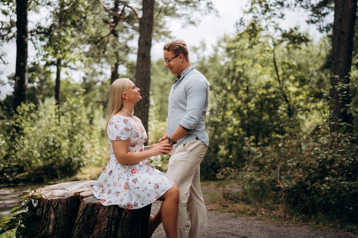 Parfotografering av et par som holder hender og kikker på hverandre i en solbelyst skogssti under en utendørs fotosesjon.