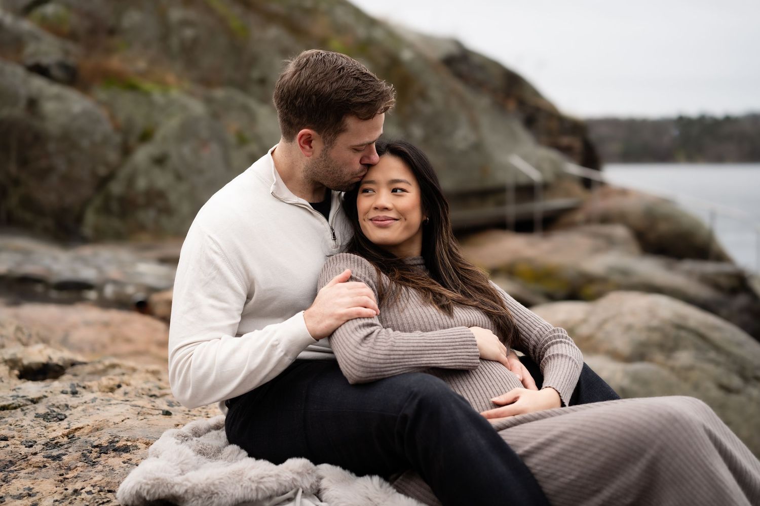 Parfotografering av et par sitter sammen langs en steinete kystlinje på en overskyet dag. Kvinnen holder over sin gravidmage.