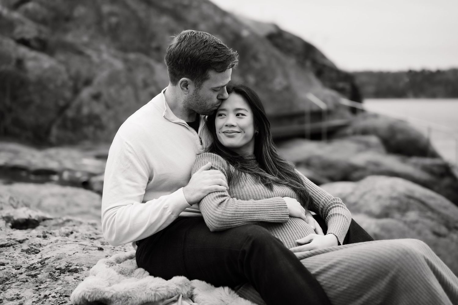 Romantisk parfotografering i svart-hvitt av et par som omfavner hverandre på steinete kystterreng. Damen er gravid.