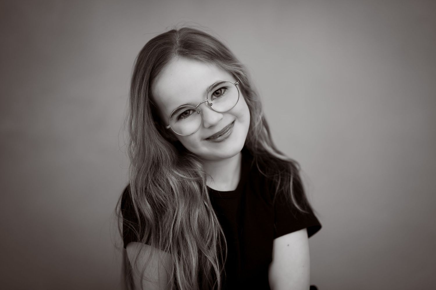 Portrettfotografering i svart-hvitt som viser ei smilende jente med briller og mørke klær.