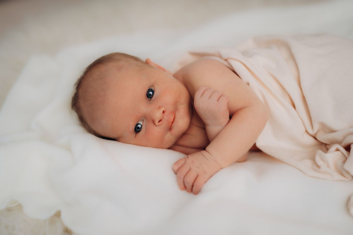 Nyfødtfotografering. En nyfødt baby ligger våken på et hvitt teppe og kikker mot kamera.
