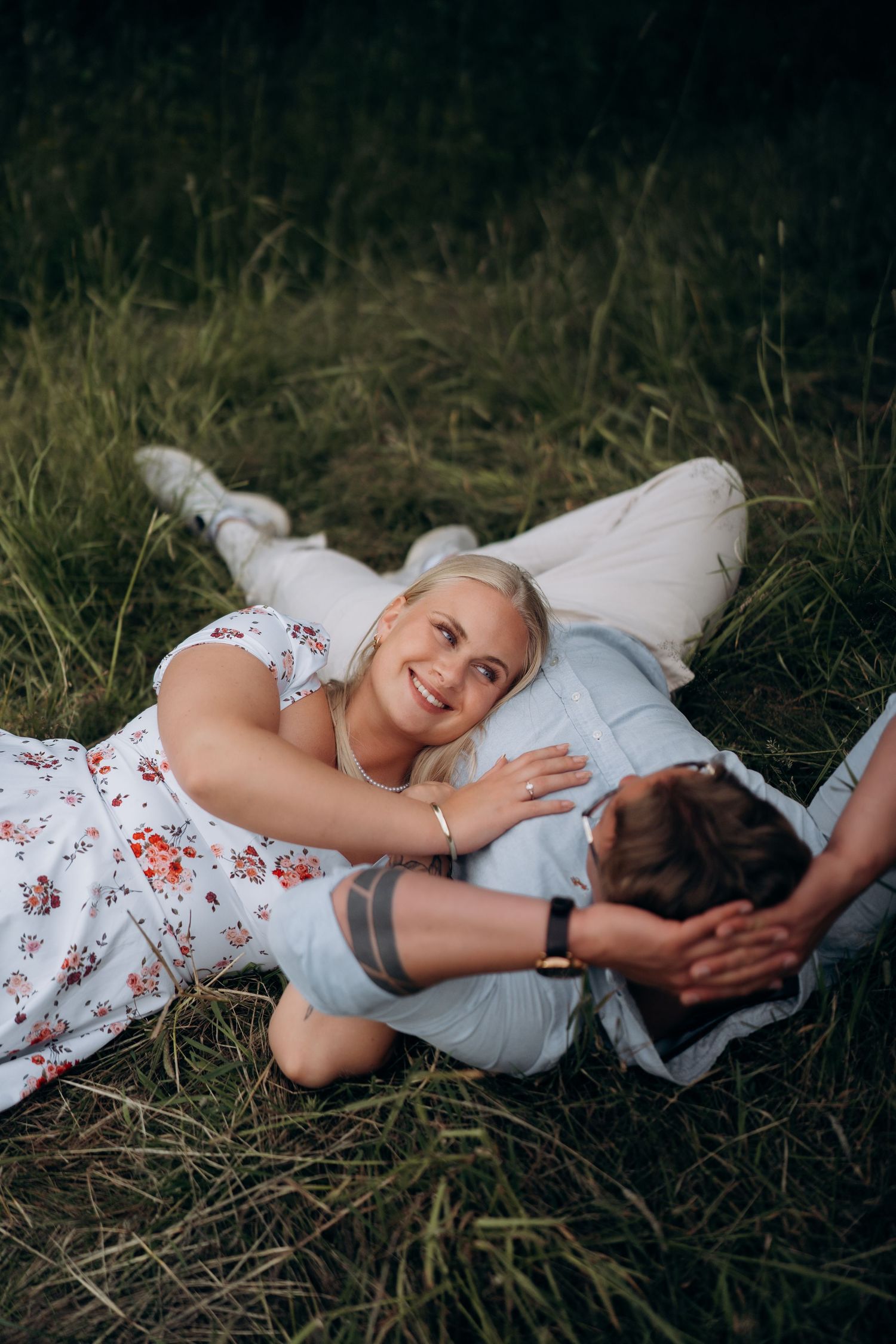 Parfotografering av et par i hvite og blomstrete antrekk som ligger sammen i gresset under en romantisk utendørs fotosesjon.