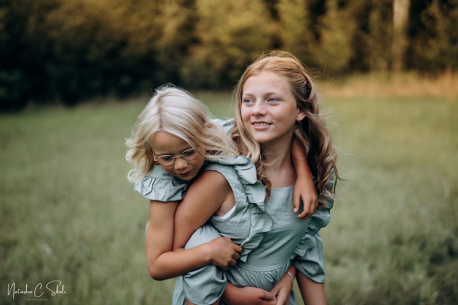 Søskenfotografering - Et smilende søskenpar i en eng ute om sommeren. Lillesøster sitter på ryggen til storesøster.