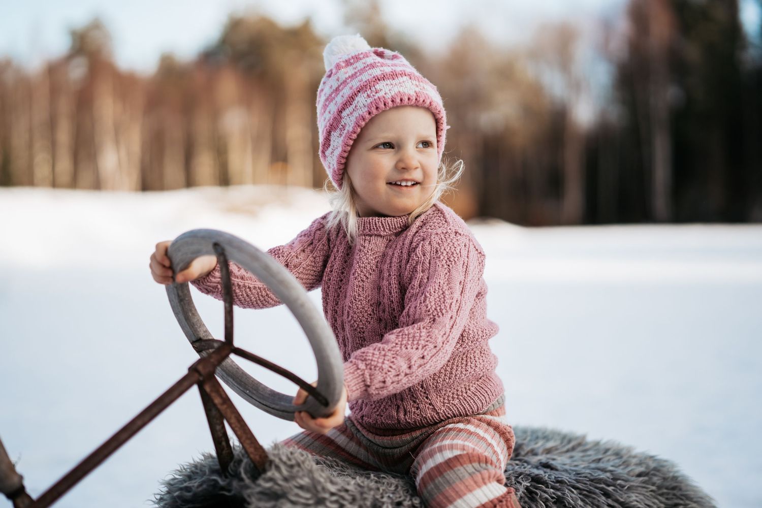 Et barnefoto av ei 4 år gammel jente som sitter på en kjelke i rosa strikkede klær.