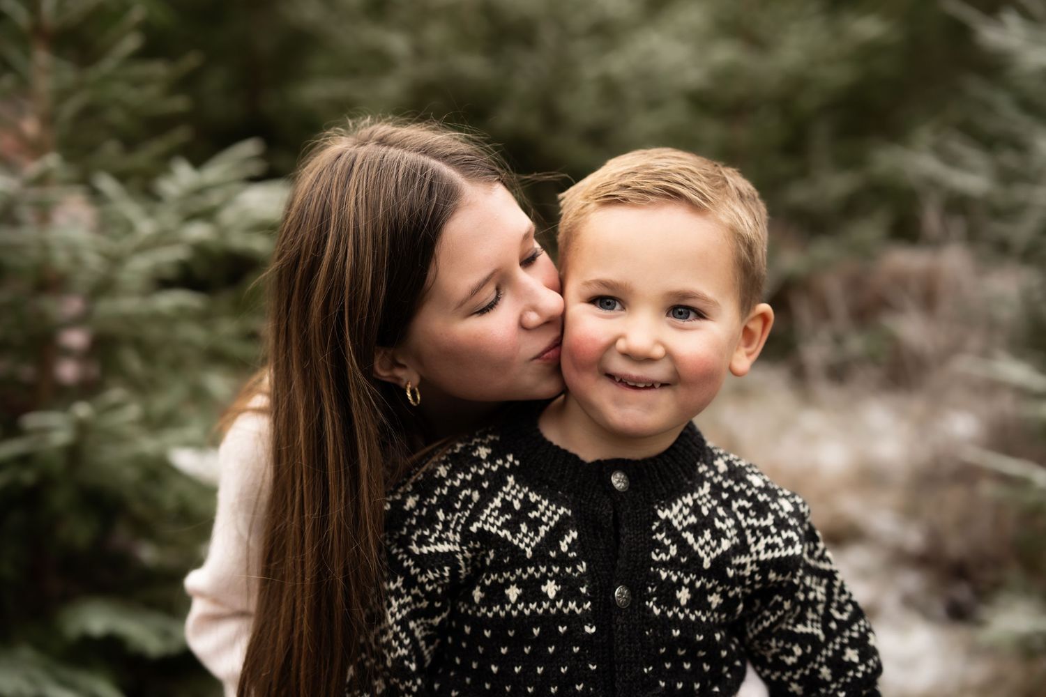 Søskenfotografering av en storesøster som kysser lillebror på kinnet. Det er vinter og begge går i varme klær i skogen.