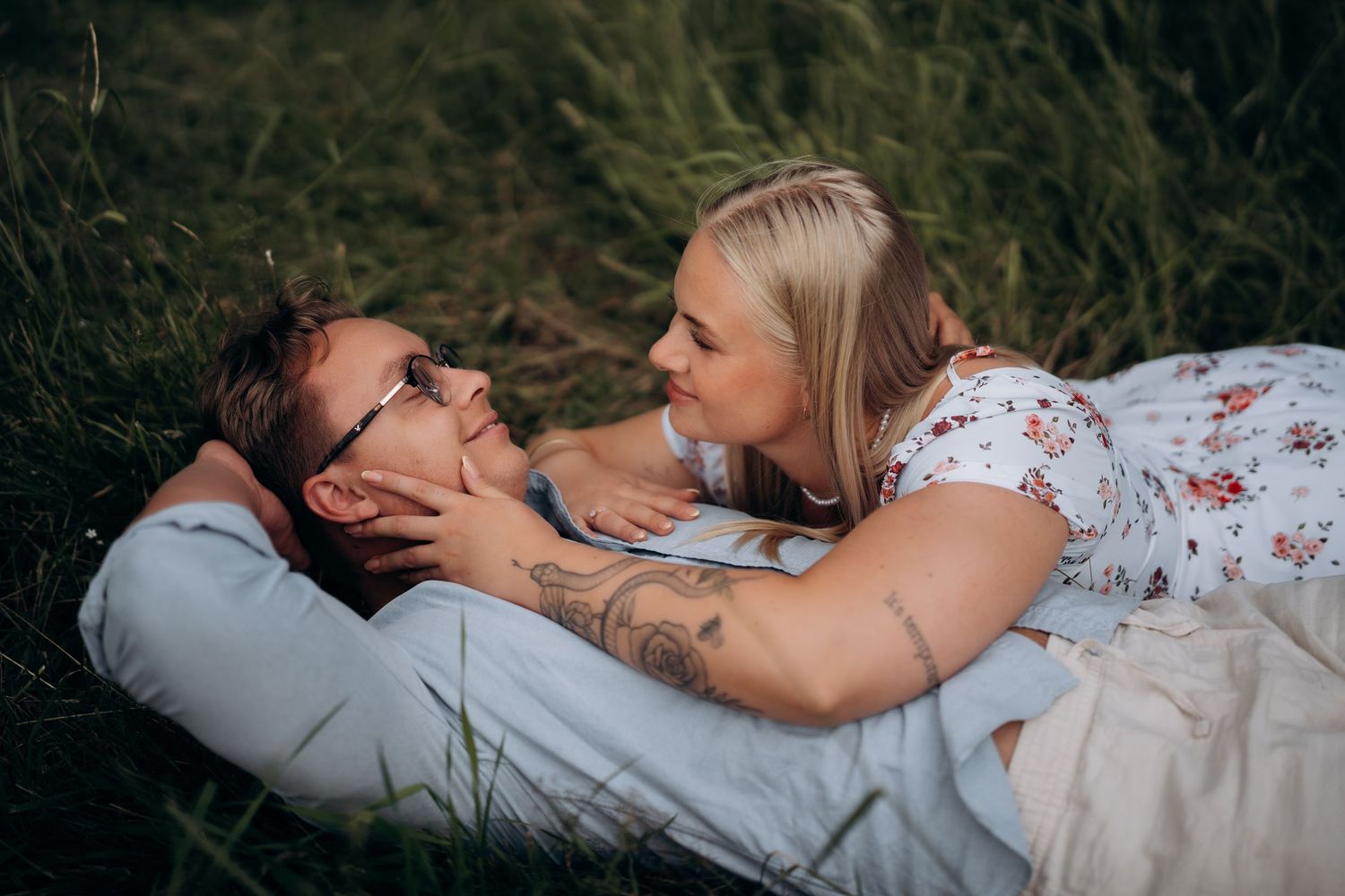 Parfotografering av et par som ligger i høyt gress og kikker på hverandre under en romantisk utendørs fotosesjon.