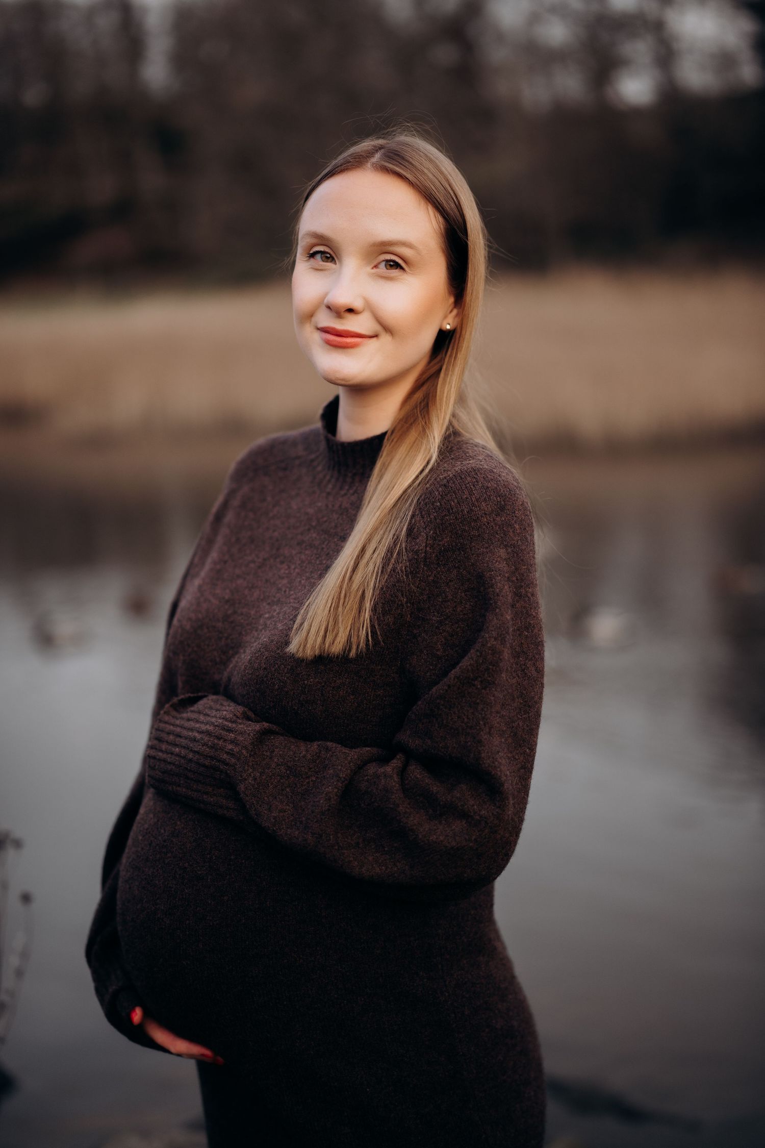 Portrett fotografering av gravid dame - damen står ute foran en innsjø og kikker smilende inn i kameraet.