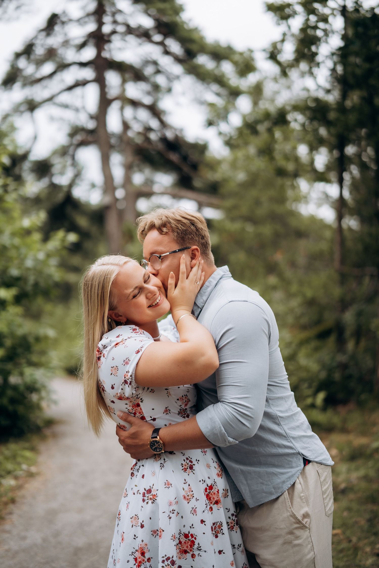 Parfotografering av et romantisk par som omfavner hverandre på en allé omgitt av trær under en utendørs fotosesjon.