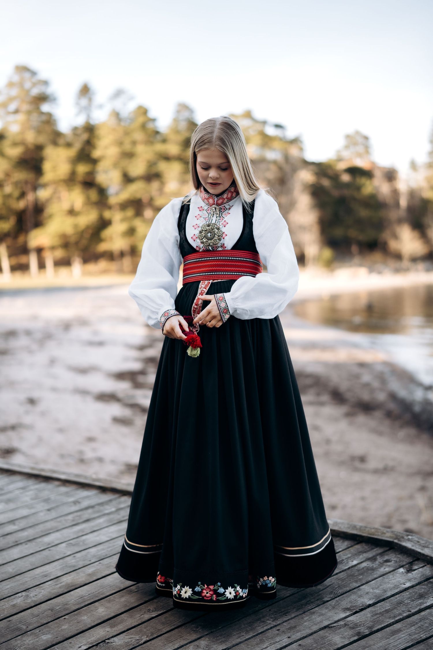 Konfirmasjonsfotografering av ei jente i som står utendørs ved vannet og retter på bunaden sin.