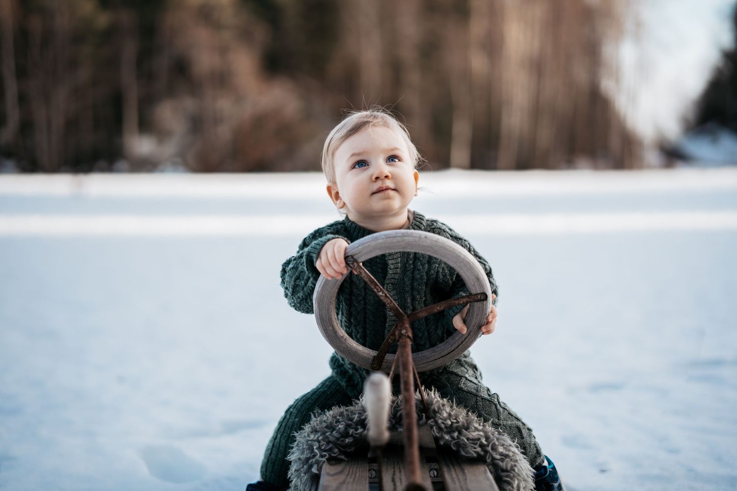 Barnefotografi av en liten gutt som sitter på en kjelke i mørkegrønn strikket dress.