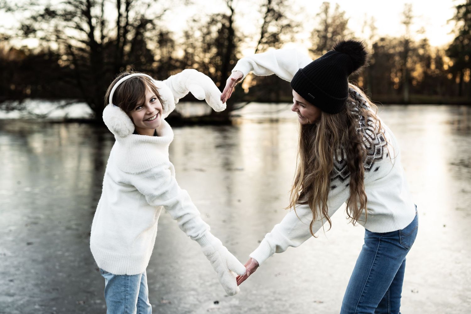 Mor og datter fotografering. Det er vinter, begge står i lyse klær foran en innsjø og lager et hjerte med hendene sine.