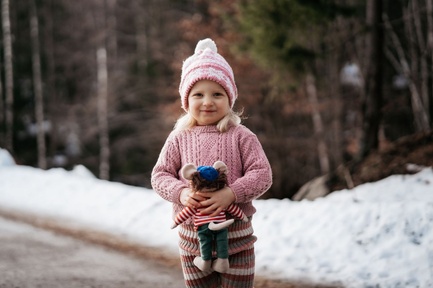 Barnefoto av ei 4 år gammel jente som står ute i snøen med bamsen sin i hånden mens hun smiler.