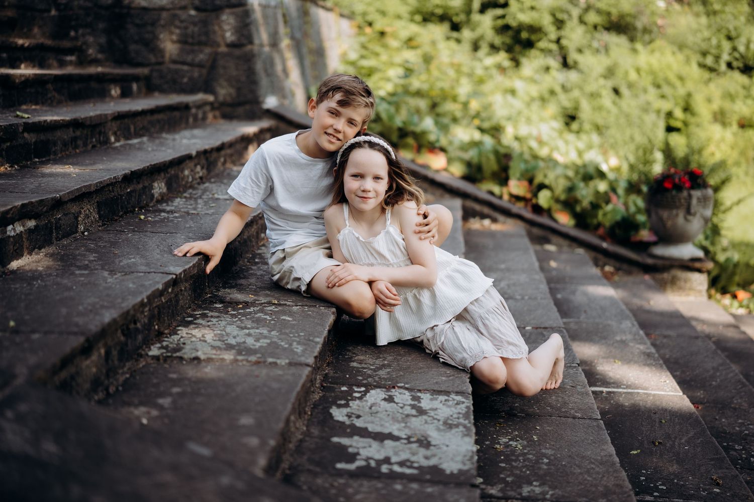 Søskenfotografering av to barn i lyse antrekk som sitter sammen på en steintrapp og smiler mot kamera.