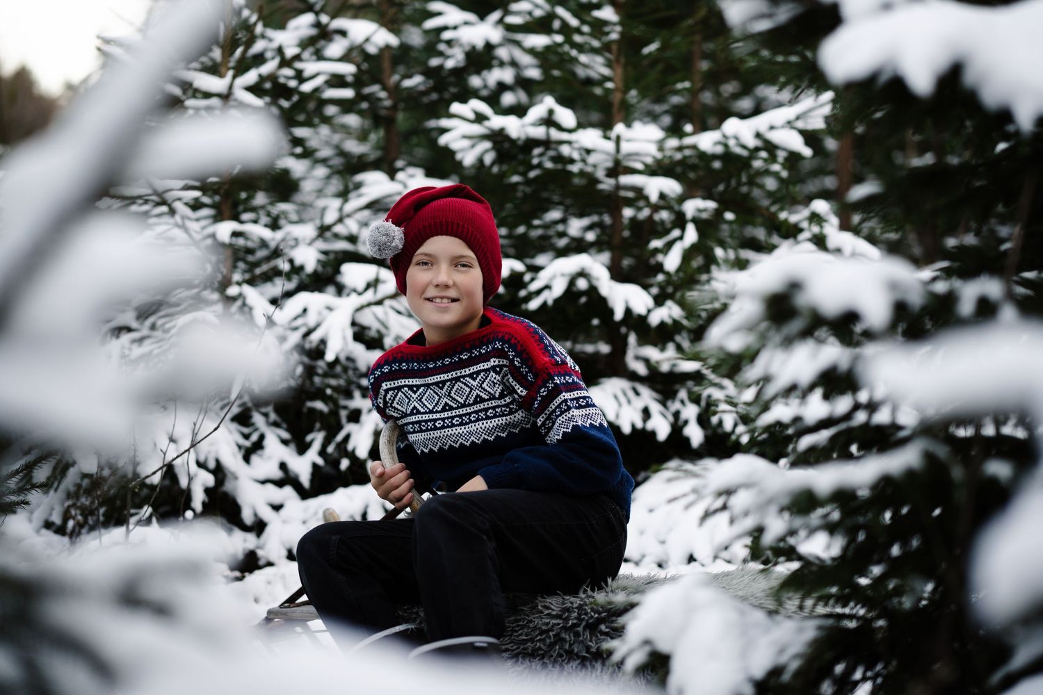 Julefoto av en gutt i mariusgenser med nisselue som. sitter på en kjelke i skogen dekket av snø.