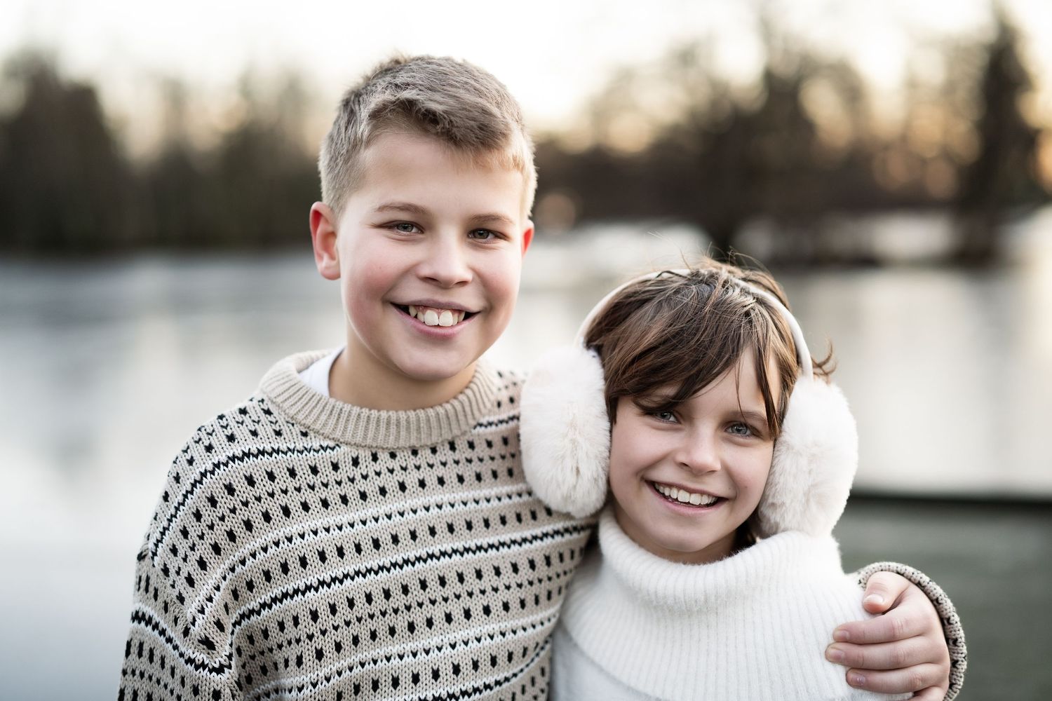 Barnefotografering av to barn i vintergensere som smiler sammen ved en innsjø under gyllent kveldslys.