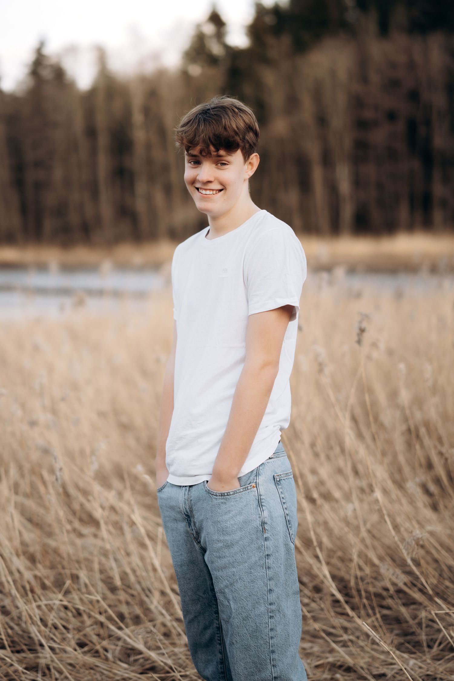 Fotografering av en ungdomsgutt som står i en åker og smiler mot kamera. Har på seg hvit t-skjorte og blå jeans.