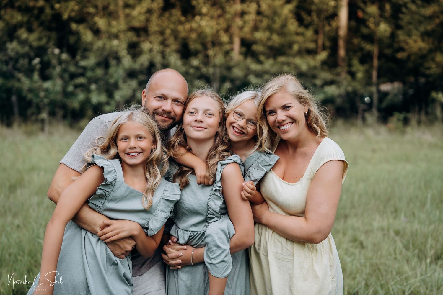 Familiefotografering - en smilende familie som står og smiler mot kamera - på en eng en sommerkveld. Mor, far og tre døtre.